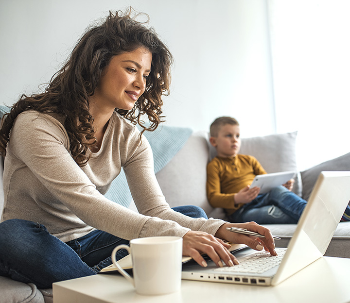 A woman working on a laptop at home while a boy uses a tablet on the sofa behind her